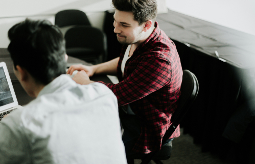 Two men sitting at a table, one using a laptop, in a casual setting.