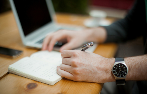 Person writing in a notebook with a laptop nearby on a wooden table.