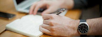 Person writing in a notebook beside a laptop and smartphone.