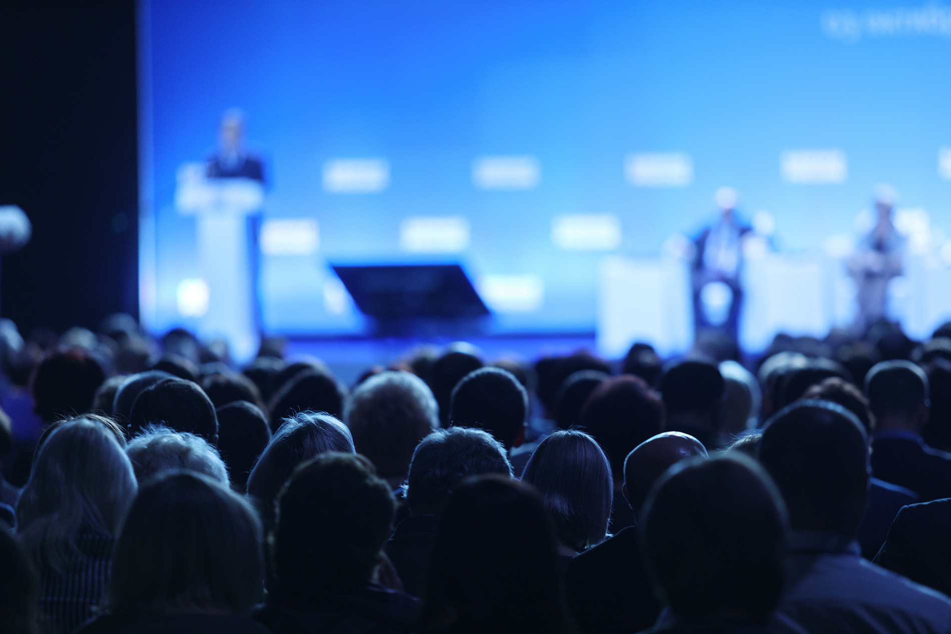 Audience in a conference hall watching speakers on a blue-lit stage.