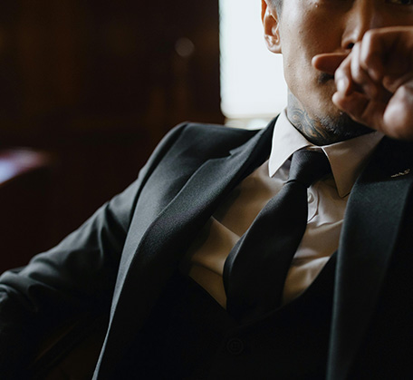 Man in a black suit and tie, sitting thoughtfully in a dimly lit setting.